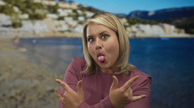 Sarışın kadın dilinden sarkan ve deniz kıyısında parlak güneş ışığı altında kaya boynuzları yapan heyecan.