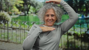 Gri saçlı bir kadın, güneşli park ortamında elleri ile, yemyeşil bir zemin üzerinde, günlük gri bir gömlek giyerek, neşe ve yaratıcılık ifade eden hareketler yapar..