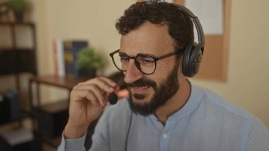 Ofis ortamında kulaklık takarken gülümseyen sakallı İspanyol bir adam profesyonel bir iş ortamı öneriyor..