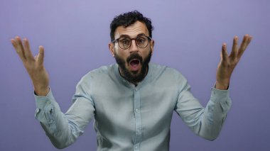 Mor bir arka plana karşı çılgın ve karışık bir ifade sergileyen sakallı İspanyol bir adam, gözlük takıyor ve açık mavi gömlek giyiyor..