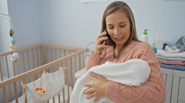 Modern yatak odasında beşiği olan bir kadın telefonda konuşuyor, rahat bir ev ortamında anneliğe ve aile dinamiklerine odaklanıyor..