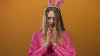 Tavşan kulaklı kadın turuncu bir arka plana gülümsüyor pembe bir gömlek giyiyor, neşeli bir paskalya temasını canlı bir şekilde yakalıyor..