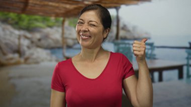 Kadın, güneşli kumsal güvertesinde turkuaz deniz manzarası ve Rocky Cove arka planıyla baş parmak hareketi gösteriyor. Onaylandı..