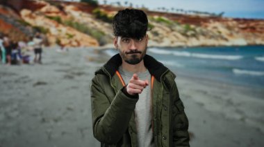 Genç İspanyol bir adam, sakin bir plaj ve okyanus arkaplanı ile düşünceli bir şekilde deniz kenarında el kol hareketi yapıyor, tefekkür ediyor ve bir dış mekâna odaklanıyor..