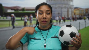 Turkuaz forma giyen gülümseyen hakem kadın futbol topunu kalabalık sokaktaki tarihi kulede tutuyor..