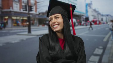 Mezuniyet şapkalı genç İspanyol kadın şehir sokaklarında gülümsüyor, açık şehir ortamındaki başarısını kutluyor..