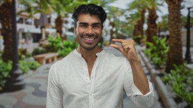 Adam, palmiye ağaçları ve bankları olan yeşil bir parkta başparmak ve işaret parmağı çimdikler; hassasiyet ölçümü.