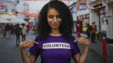 Latin bir kadın, yardım kampanyası sırasında şehir caddesinde baş parmağıyla mor gönüllü gömleğini işaret ediyor..