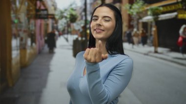 Mavi tişörtlü genç İspanyol kadın şehir caddesinde neşeli bir ifade sergileyerek hareketli bir jest yapıyor.