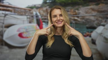 İki eliyle gülümseyen ve yanaklarını gösteren bir kadın güneşli bir günde arka planda teknelerle kumlu bir sahilde duruyor..