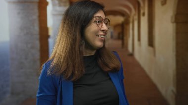 Mavi ceketli ve gözlüklü kadın, güneşli bina koridorunda başını çevirirken gülümsüyor; yolculuk keşfi huzur.