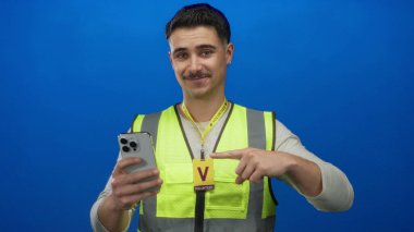Yansıtıcı yelek giymiş genç İspanyol bir adam, akıllı telefonuyla, modern teknoloji ve gönüllülüğü somutlaştıran, mavi izole bir arka plana işaret ediyor..
