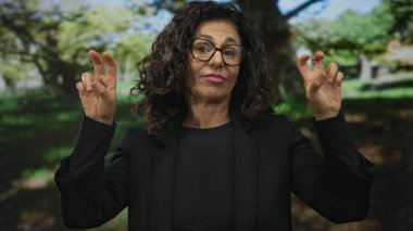 Orta yaşlı İspanyol kadın, güneşli yeşil bir parkta parmak kaldırarak tırnak işareti yapıyor; iğneleme şüpheciliği.