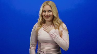 Elini göğsüne koyup gülümseyen bir kadın canlı mavi arka planda minnettarlığını ve pozitifliğini ifade ediyor..