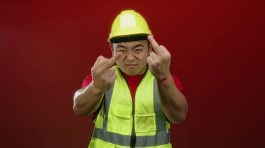 İnşaat işçisi koyu kırmızı arka planda anlamlı bir jest yapıyor sarı kaskını ve yansıtıcı yeleğini vurguluyor..