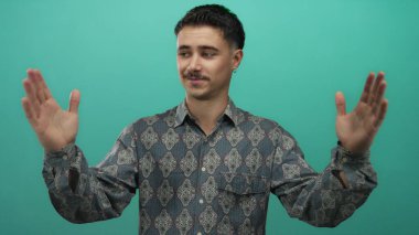 Genç İspanyol adam yeşil arka plan duvarının önünde elleri ile büyük jest yapıyor, desenli gömlek ve küpeler giyiyor, boyunu veya mesafesini ifade ediyor..