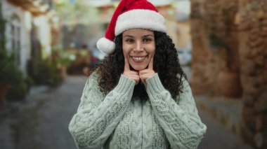 Noel Baba şapkalı bir kadın açık hava sahnesinde gülümsüyor ve neşeli bir ifadeyle bayram sezonunu kutluyor..