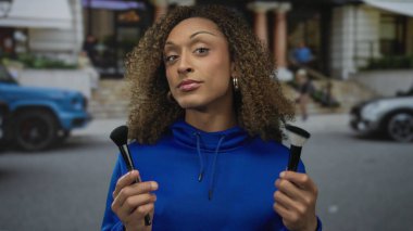 Arkasında bulanık bir bina cephesi olan iki makyaj fırçasını yakından inceleyen kadın, belirsizlik.