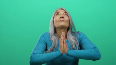 Dua eden gri saçlı yaşlı kadın, parlak yeşil bir arka plana karşı poz veriyor, mavi bir kazak giyiyor ve sakin, ruhani ve bilinçli bir şekilde izole edilmiş stüdyo ortamından geçiyor..