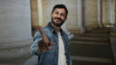 Sakallı bir adam Roma 'daki antik sütunların arasında jest yapmaz, tarihi eski şehir ortamında dinamik bir anı yakalar kot ceket ve kendine güvenen bir ifadeyle.