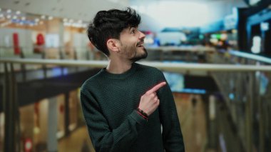 Genç adam, hareketli bir alışveriş merkezini işaret ederek canlı bir alışveriş havası ve şaşkınlık ve merak ifadeleri sergiliyor..