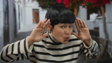 Şehir sokaklarında gülen çiçeklerin altında çizgili süveterle gülümseyen neşeli dış görünüşlü bir kadın şehir ortamında neşe ve doğallığın yakalandığını gösteriyor..