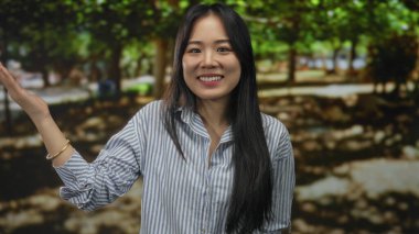 Yeşil bir parkta arka planda ağaçlarla gülümseyen, çizgili bir gömlek giyen, açık havada mutluluk ve neşe saçan genç Çinli kadın..