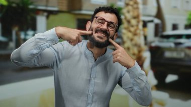 Sakallı İspanyol bir adam, şehir ortamında mizah ve ifadeyi vurgulayan bulanık bir sokak geçmişi olan sıradan bir tişörtle dışarıda komik suratlar yapıyor..