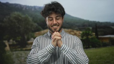 Yeşil bir parkta gülümseyen sakallı genç adam rahat ve mutlu bir tavır sergiliyor çizgili bir gömlek giyiyor yemyeşil ağaçlar ve dingin dağ manzarasına karşı.