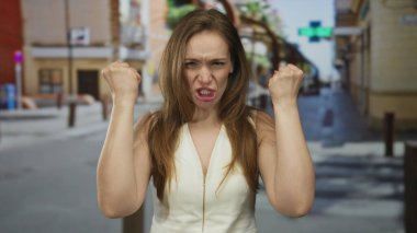 Beyaz kolsuz üst kollu bir kadın, güneşli bir şehir sokağında dükkanlarla dolu yumruklarını ve hırıltılarını sıkıyor..