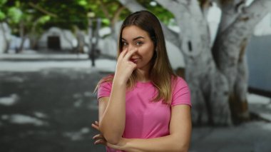 Pembe tişörtlü bir kadın, güneşli bir günde, açık havada, şehir sokağındaki bir ağacın altında neşeli bir ifade sergiliyor..