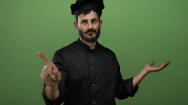 Siyah üniformalı İspanyol erkek aşçı, yeşil bir zemine parmağıyla hayır işareti yapıyor. Mutfak bağlamında onaylamıyor..