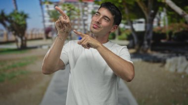 Gülümseyen İspanyol adam, parlak güneş ışığı altındaki yeşil bir parkta iki eliyle objeyi işaret ediyor..