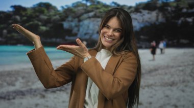 Genç İspanyol kadın, iki eliyle sahilin manzaralı sahnesinde turkuaz deniz ve arka planda bulanık ağaçlarla bir şey sunuyor..