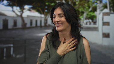 Orta yaşlı İspanyol kadın göğsünü tutuyor. Yüzüklü parmaklarıyla yeşil yaprakların altında gülüyor. Minnettar sevinç..