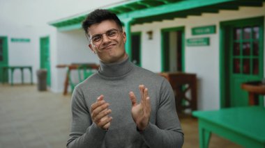 Genç bir adam şehir restoranının terasında neşeli bir şekilde alkışlıyor, gündelik ortamında çekici ve mutlu bir adamı sergiliyor..