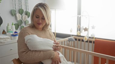 Stüdyoda ahşap beşiğin yanında beyaz battaniyeye sarılı uyuyan bebeği nazikçe kucaklar; anne sevgisi dinginliği.