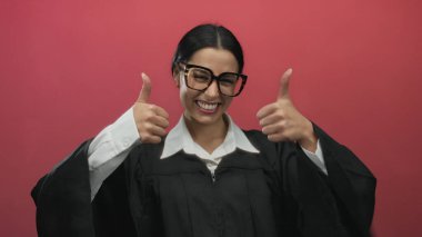 Siyah cüppeli, gözlüklü bir kadın gülümsüyor ve canlı kırmızı arka plana başparmak sallıyor. Kendine güvenen ve profesyonel tavırlarını dışa vuruyor..