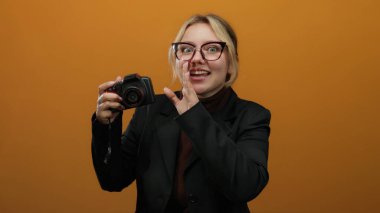 Bir kadın gizlice kamerayı turuncu duvara dayamış, gözlüklü ve siyah ceketli, sarı saçlı, şaşırmış, izole edilmiş, hayat dolu arka plan..