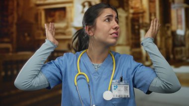 Mavi önlüklü ve kimlik kartlı genç doktor, kilisenin iç kısımlarında ahşap sıraların arasında sarı steteskopla kulakları örtüyor..