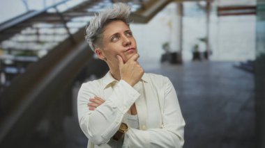 Merdivenin yanındaki bina lobisinde eli çenesinde düşünceli bir kadın kol saati takıyor; iç yansıması.