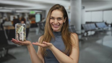 Elinde yuvarlanmış banknotlarla dolu cam bir kavanoz tutan ve yoğun bir havaalanı terminalinde onu işaret eden gülümseyen kadın; gelecekteki iyimserlik.