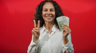 Beyaz gömlekli orta yaşlı bir kadın canlı kırmızı bir duvara karşı Şili pezosu taşıyor. Bu da izole bir ortamda başarı ve finans simgesi..