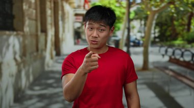 Kırmızı tişörtlü genç adam, güneşli bir şehir sokağında bir telefon konuşması yapıyor ve şehir ortamında iletişim kurduğunu söylüyor..