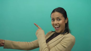 Bej renkli gülümseyen İspanyol kadın enerjik bir şekilde yeşil bir arkaplanı işaret ediyor, heyecan ve pozitifliği ifade ediyor..