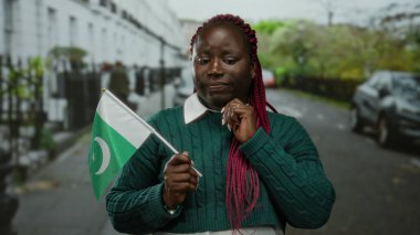Bir şehir caddesinde elinde pakistan bayrağı olan bir kadın düşünceli görünüyor, kültürel çeşitliliği şehir içi araba ve binalarla harmanlıyor..