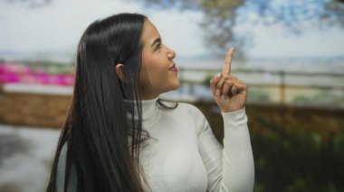 Beyaz kazaklı kadın, açık havada gülümseyen arka planda bulanık bir gülümseme ile yukarı bakıyor. Çiçekler ve yeşillik ile dolu bahar ya da yaz sezonunu öneriyor..