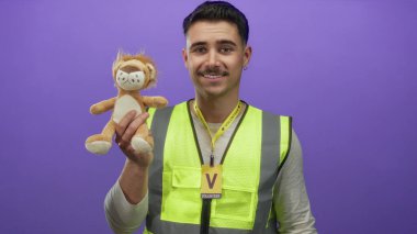Yansıtıcı yelek giymiş genç İspanyol bir adam izole edilmiş mor arka plana karşı oyuncak bir aslan tutuyor gülümseyerek gönüllü oluyor.