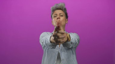 Kısa gri saçlı, çizgili tişörtlü bir kadın stüdyo ortamında iki eliyle parmak tabancası hareketi yapıyor..