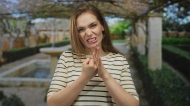 Avucunun içine sıkılmış bir yumruğu sıkıştıran kadın, güneşli bir parkta çitlerle ve taş bir çeşmeyle kaşlarını çatarken; öfke..